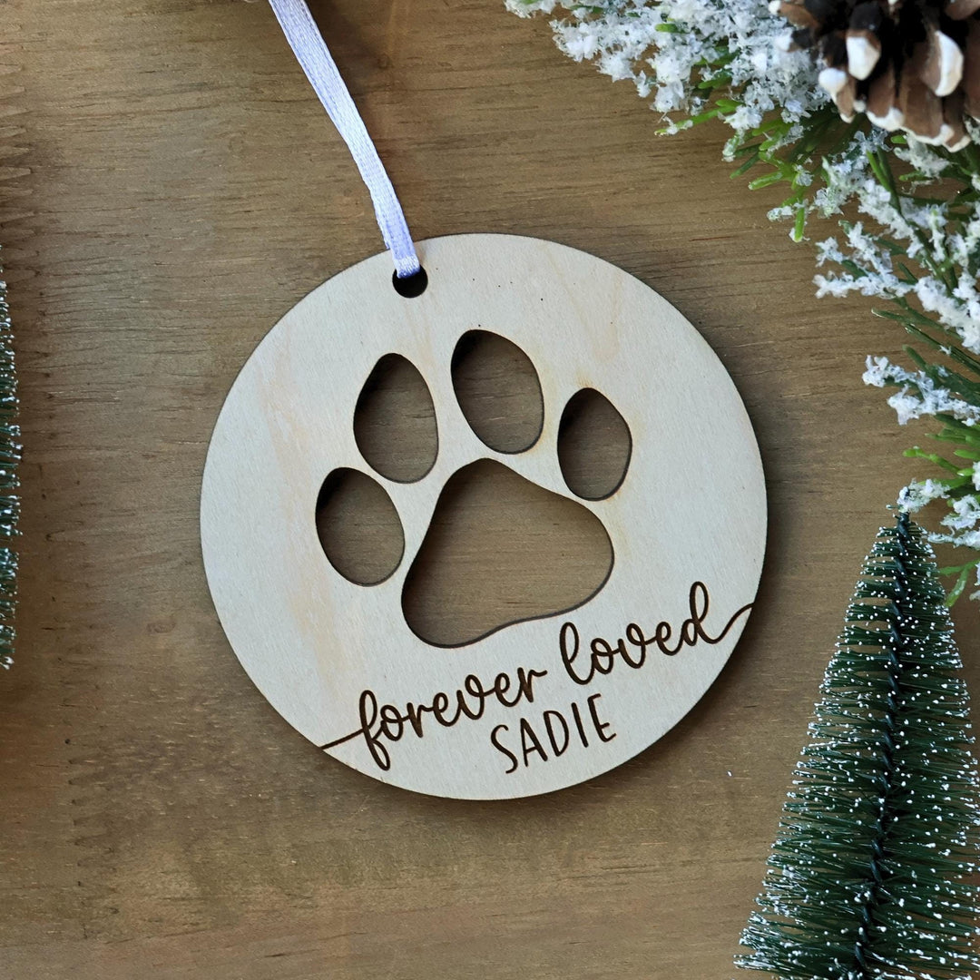May include: A round wooden Christmas ornament features a cutout paw print and the engraved text, 'forever loved Sadie.' The ornament is light beige and has a white ribbon attached.  It's a pet memorial ornament, perfect for remembering a beloved pet. The design is simple and elegant, suitable for a pet loss keepsake.